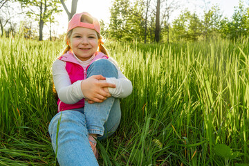 A cheerful girl sits in the thick green grass, clasping one leg with her hand, against the backdrop of the sunset. Walk outdoors in the city park