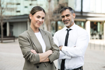 Confident and smiling business people looking at camera in the street with their arms crossed. Young businesswoman with a senior businessman behind looking satisfied.