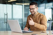 © Tetiana - A smiling man sits at a table in an office center, works at a laptop and talks via video call, business training, online conference with partners, clients, project discussion.
