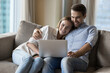 Positive young husband and wife resting on sofa, using laptop for Internet communication, enjoying wireless technology, online service, leisure hugging, smiling, watching movie at home