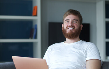 Wall Mural - Young happy positive man use laptop computer at home in living room sit on couch looking at camera and smile. Freelancer guy, freelance distant job