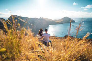 Wall Mural - Back view of a tourist couple sitting on a field with coastal landscape of Madeira Island in the Atlantic Ocean in the morning. São Lourenço, Madeira Island, Portugal, Europe.