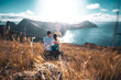 © Michael - Back view of a tourist couple sitting on a hill, overlooking the coastal landscape of Madeira Island in the Atlantic Ocean in the morning. São Lourenço, Madeira Island, Portugal, Europe.