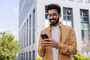 Successful Indian young businessman outside office building walking in daytime, man holding phone in hands, businessman dialing, browsing online pages, programmer engineer in glasses.