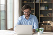 © fizkes - Positive handsome freelancer businessman in glasses working at home office table, typing on laptop, using wireless technology for Internet job communication, smiling. Candid front portrait