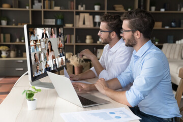 Focused twin family business partners speaking on online conference video call, using large desktop monitor with head shots, talking to diverse freelance team of colleagues, discussing teamwork