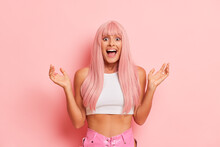 Waist-length Photo Of Happy Girl With Long Pink Hair In White Top Stands Isolated On Pink Backdrop Hands Up With Joy, Happy Time Concept, Copy Space