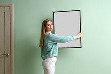 Young Woman Hanging Blank Frame On Green Wall At Home