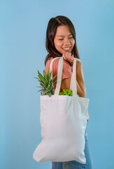 Wall Mural - happy young asian housewife carrying reusable cloth bag of fruits and vegetables isolated on blue background,concept for campaign of using reusable bag, reduce waste,stop global warming,save the world