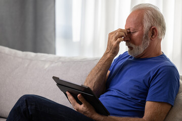 Senior man using digital tablet computer at home and having eyestrain or suffering from astigmatism and bad vision. Eyesight problems concept.