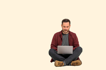 Wall Mural - Focused handsome businessman dressed in maroon shirt working over laptop and sitting with crossed legs. Young busy male entrepreneur analyzing financial data on wireless computer against background