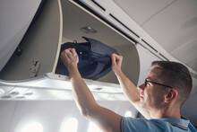 Man travel by airplane. Passenger putting hand baggage in lockers above seats of plane..