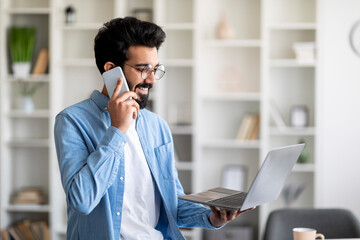 Wall Mural - Remote Business. Indian Man Talking On Cellphone And Holding Laptop At Home