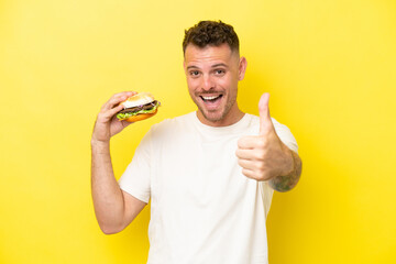 Wall Mural - Young caucasian man holding a burger isolated on yellow background with thumbs up because something good has happened