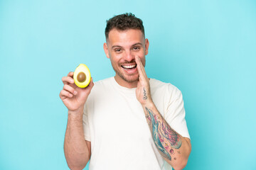 Wall Mural - Young caucasian man holding an avocado isolated on blue background shouting with mouth wide open