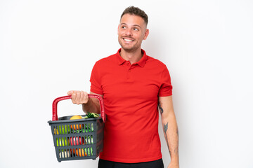 Wall Mural - Young caucasian man holding a shopping basket full of food isolated on white background thinking an idea while looking up