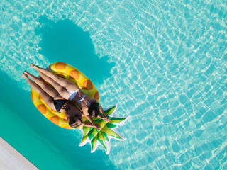 Wall Mural - Top view of two girls relaxing on inflatable big pineapple in the pool. Aerial view of ladies relaxing on the floating mattress.