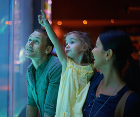 Wall Mural - Family, aquarium and child pointing at fish for learning, curiosity or knowledge, bonding or care. Mother, fishtank and happy kid with father watching marine animals swim underwater in oceanarium.