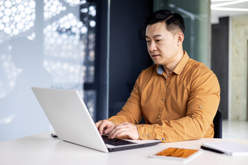 serious and focused businessman working with laptop, man in glasses and shirt inside office building