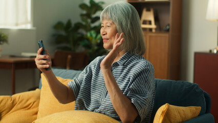 Wall Mural - Happy attractive Asian elderly woman using smartphone video conference call in cozy living room. Happy old aged female using mobile phone technology chatting talking with family or friends at home