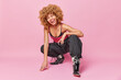 Full length shot of fashionable curly woman squats against pink background winks eye and sticks out tongue wears stylish clothing isolated over pink backgroun has flirty expression. Fashion model