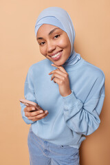 Wall Mural - People and gadgets concept. Smiling Islamic woman touches chin gently holds mobile phone reads messages and browses internet wears hijab pullover and velvet trousers isolated over brown background