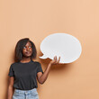 © wayhome.studio  - Pensive dark skinned young woman has thoughtful expression holds empty speech bubble thinks which information to place there dressed in black t shirt and jeans isolated over brown background