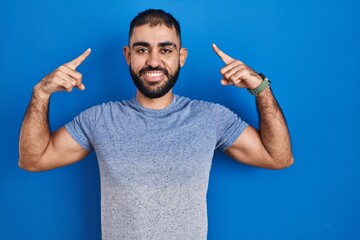 Sticker - Middle east man with beard standing over blue background smiling pointing to head with both hands finger, great idea or thought, good memory