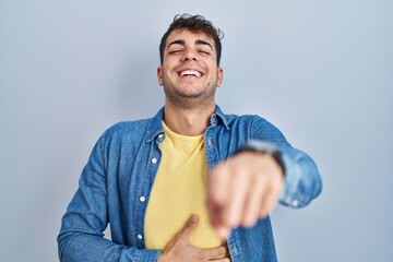 Sticker - Young hispanic man standing over blue background laughing at you, pointing finger to the camera with hand over body, shame expression