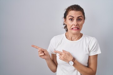 Sticker - Beautiful brunette woman standing over isolated background pointing aside worried and nervous with both hands, concerned and surprised expression