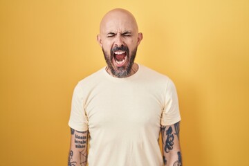 Wall Mural - Hispanic man with tattoos standing over yellow background angry and mad screaming frustrated and furious, shouting with anger. rage and aggressive concept.