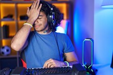 Hispanic Man With Curly Hair Playing Video Games Surprised With Hand On Head For Mistake, Remember Error. Forgot, Bad Memory Concept.