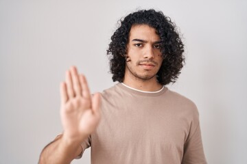 Wall Mural - Hispanic man with curly hair standing over white background doing stop sing with palm of the hand. warning expression with negative and serious gesture on the face.