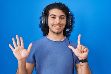 Poster - Hispanic man with curly hair listening to music using headphones showing and pointing up with fingers number seven while smiling confident and happy.