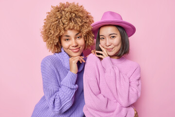 Wall Mural - Portrait of two female friends stand closely to each other have natural gentle smile wear knitted jumpers hat isolated over pink wall meet together at weekend. Beautiful women in fashionable clothes