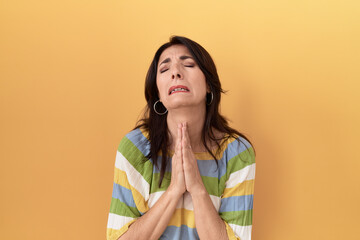 Middle age hispanic woman standing over yellow background begging and praying with hands together with hope expression on face very emotional and worried. begging.