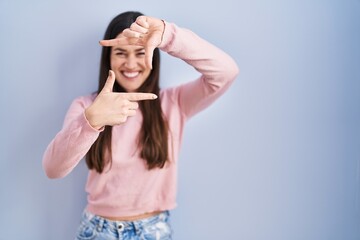 Canvas Print - Young brunette woman standing over blue background smiling making frame with hands and fingers with happy face. creativity and photography concept.