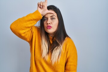 Wall Mural - Young hispanic woman standing over isolated background making fun of people with fingers on forehead doing loser gesture mocking and insulting.