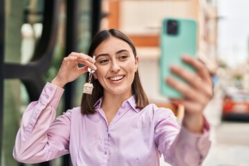 Wall Mural - Young hispanic woman make selfie by the smartphone holding key of new home at street