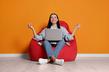 Canvas Print - Happy woman with laptop sitting on beanbag chair and meditating near orange wall