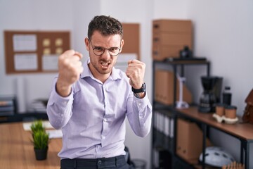 Canvas Print - Young hispanic man at the office angry and mad raising fists frustrated and furious while shouting with anger. rage and aggressive concept.