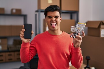 Poster - Hispanic man working at small business ecommerce holding cart sticking tongue out happy with funny expression.