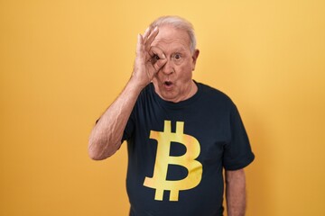 Canvas Print - Senior man with grey hair wearing bitcoin t shirt doing ok gesture shocked with surprised face, eye looking through fingers. unbelieving expression.