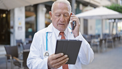 Canvas Print - Senior grey-haired man doctor using touchpad talking on smartphone at coffee shop terrace