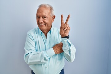 Sticker - Senior man with grey hair standing over blue background smiling with happy face winking at the camera doing victory sign. number two.