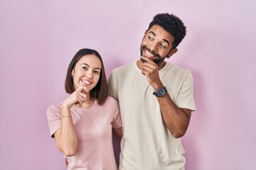 Poster - Young hispanic couple together over pink background with hand on chin thinking about question, pensive expression. smiling and thoughtful face. doubt concept.