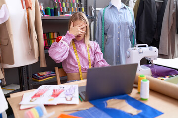 Canvas Print - Young caucasian woman dressmaker designer using laptop peeking in shock covering face and eyes with hand, looking through fingers afraid