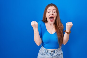 Wall Mural - Redhead woman standing over blue background excited for success with arms raised and eyes closed celebrating victory smiling. winner concept.