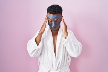 Canvas Print - Young hispanic man wearing beauty face mask and bath robe with hand on head, headache because stress. suffering migraine.