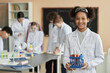 © Seventyfour - Waist up portrait of black teen schoolgirl wearing lab coat in science class and smiling at camera with group of children in background, copy space